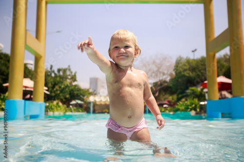 closeup small blonde girl stands in pool water points towards photo