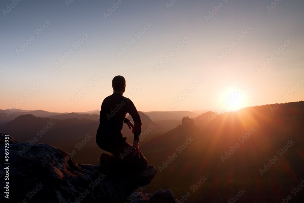 Young man in black sportswear sit on cliff edge and look into daybreak at horizon over misty valley