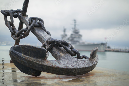 Fototapet Anchor on the embankment and the cruiser in the port of Novoross