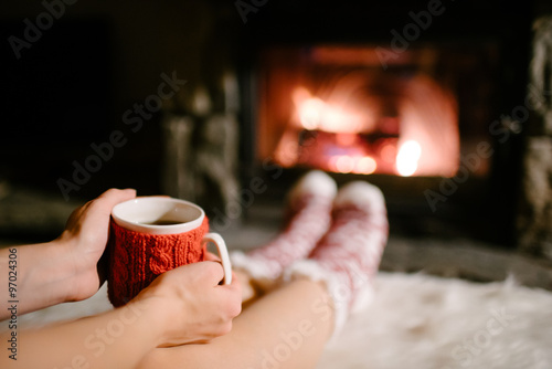 Woman holding a cup of tea by the Christmas fireplace. Woman relaxes by warm fire with cup of hot drink and warming up her feet in woollen socks. Close up on feet. Winter, Christmas holidays concept.