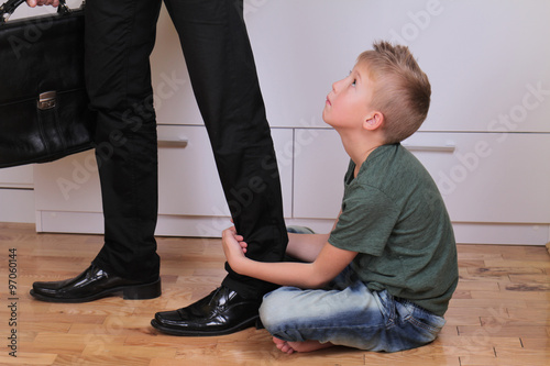 Boy holding dad's leg as he tries to leave for work. Child trying to stop father to go to work. Kid needs parent attention. Busy businessman father. Balance between work and time spent with family