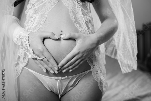 Picture closeup on bridal hands holding love heart sign on pregnant bare belly. Black and white photography