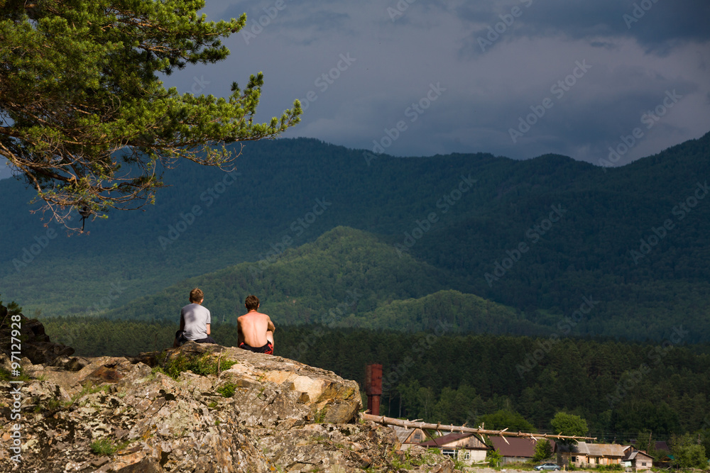 Altai village Manzherok. Katun River. Two tourist young men sitting on rocky cliff and enjoying view