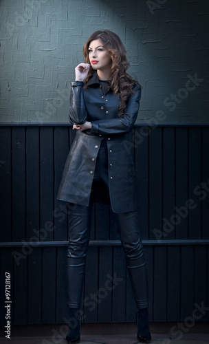 Charming young brunette woman in black leather outfit, coat and trousers, with dark gray wall on background. Sexy gorgeous young woman. Full length portrait of a sensual woman with long curly hair