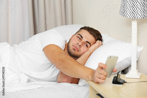 Beautiful young man waking up with mobile alarm clock photo