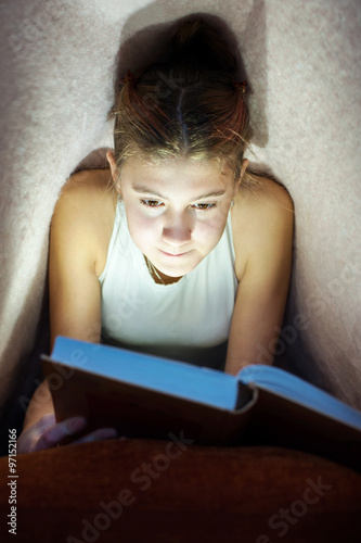 Young cheerful girl hiding under blanket and enrapt reading book photo