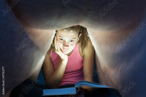 Young girl hiding under blanket and enrapt reading interesting b photo