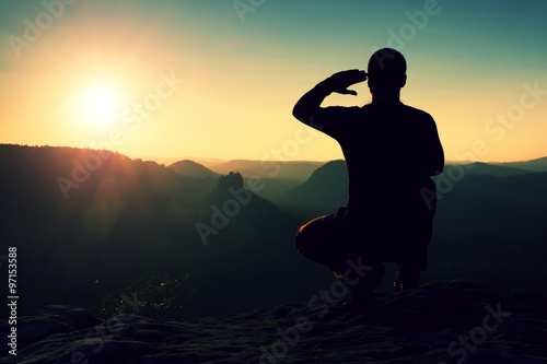Alone sportsman in black. Tall hiker in squatting position enjoy view at sunset on mountain peak