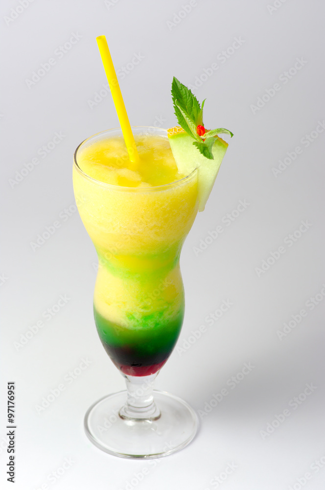 descending close-up of a hurricane glass filled with a layered fruit cocktail, with purpple and green syrup on the bottom, decorated with a melon wedge and a yellow straw, on a gray background descending close-up of a hurricane glass filled with a layered fruit cocktail, with purpple and green syrup on the bottom, decorated with a melon wedge and a yellow straw, on a gray background