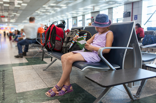 Young Caucasian girl reading book in the waiting room at the airport terminal, sitting on a chair. photo