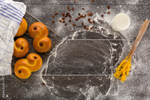 New baked swedish saffron buns, also called Lussekatter, on a floury wooden table with a glass of milk and wooden fork with sticky dough.