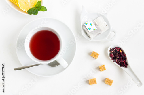 Tea set with a cup of tea, lemon, filter in teapot-shaped bowl, cube sugar and flowers. White background, top view