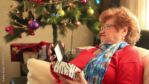 Christmas Gift -   Sulky senior woman lying on a sofa near the christmas tree,  opening gift disappointed and unhappy,