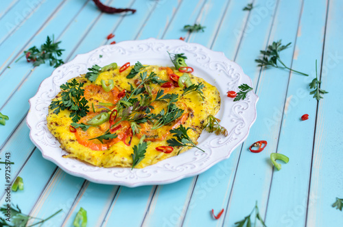 Delicious breakfast omelette or scrambled eggs with red and yellow tomatoes, red hot chili peppers and parsley on patterned plate over stylized old aged turquoise table background close-up, copy space
