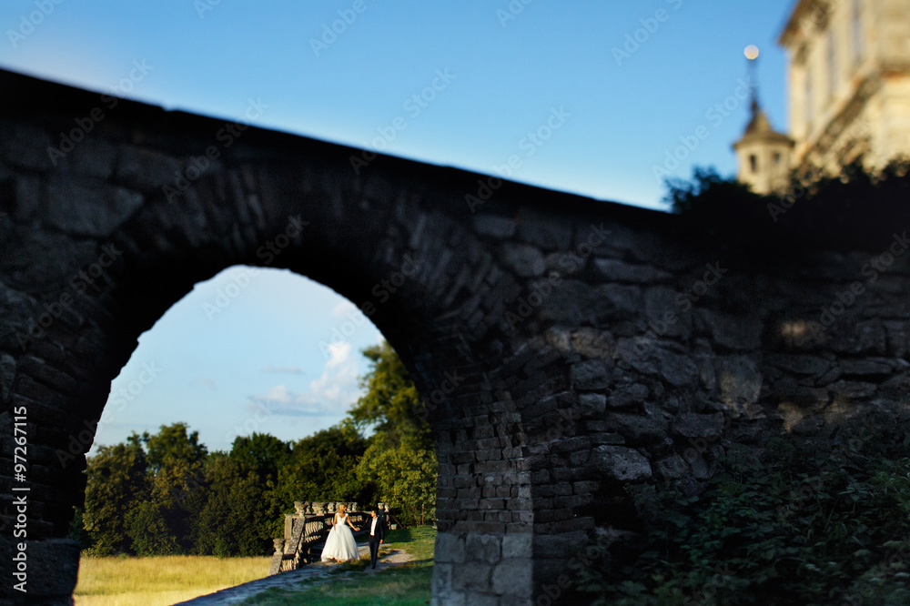 Stylish elegant bride and handsome groom holding hands walking under ancient stone bridge