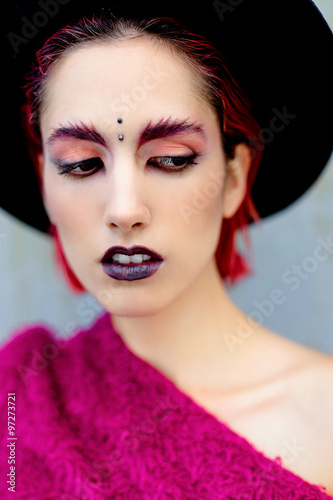 Attractive young woman with colorful makeup and piercing on forehead in black rounded hat posing with gray wall on background
