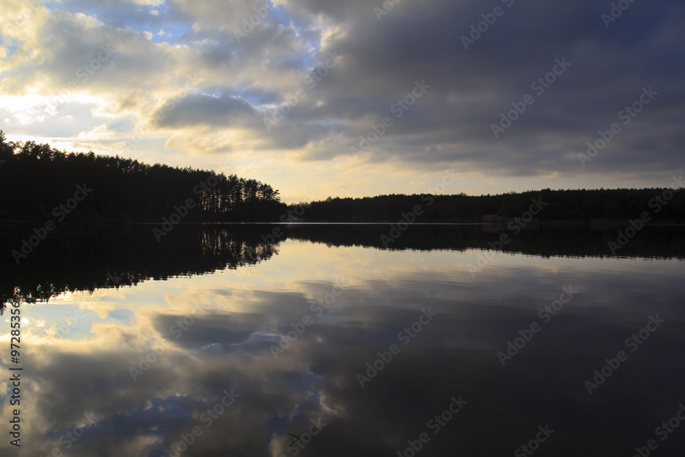 Fototapeta premium calm lake surface with morning sky reflected and dark pine forest at horizon