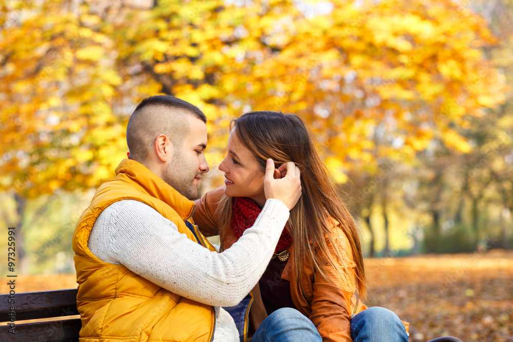 Young couple sitting on a park bench on a sunny autumn day and expressing tenderness Young couple sitting on a park bench on a sunny autumn day and expressing tenderness