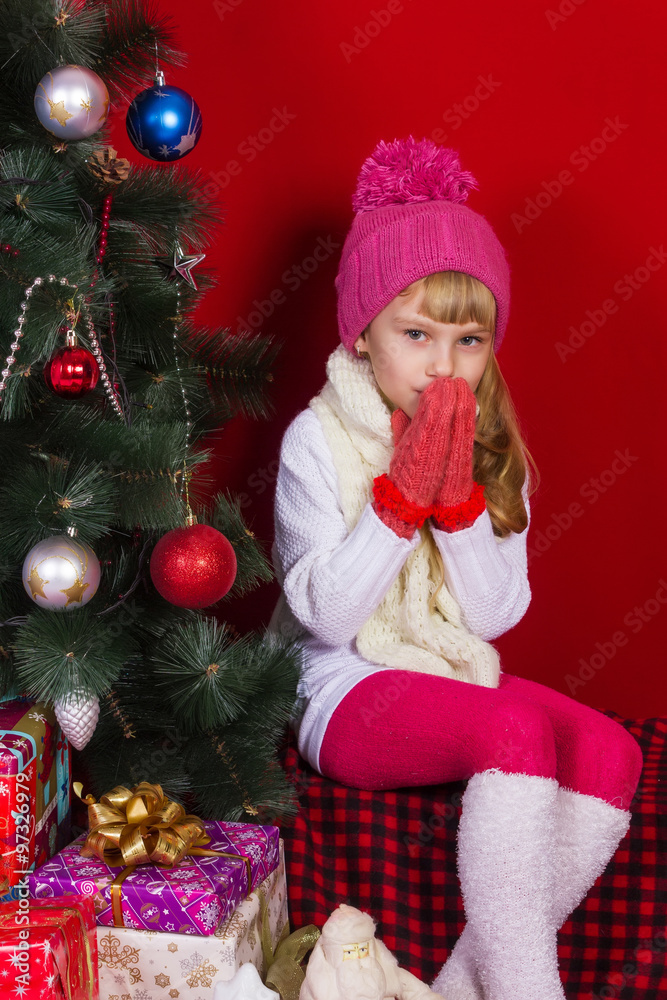 Beautiful baby girl in a pink hat and gloves near the Christmas tree in New Year's Eve smiling and looking for a gift