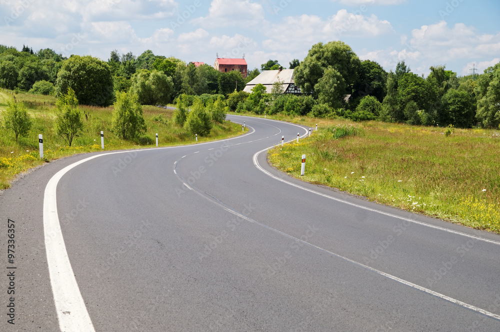 Empty asphalt road with a double bend between flower meadows in the  countryside. Village among the green leafy trees background. Clear sunny  day with blue skies and white clouds. Stock Photo |
