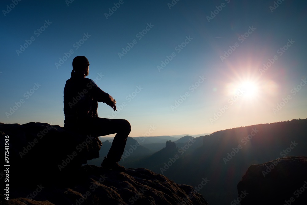 Young man in black sportswear is sitting on cliff and looking to misty valley bellow