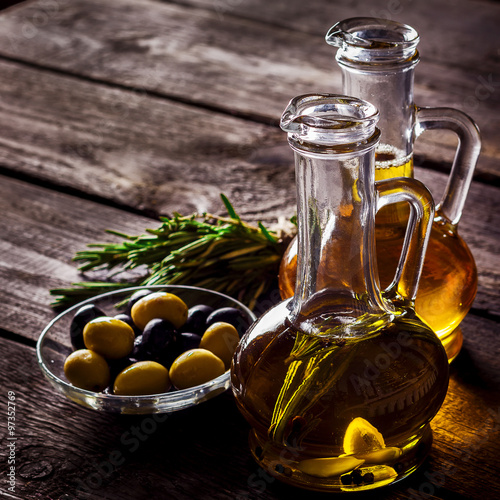 Two bottles of olive oil, olive in a bowl and herbs on a wooden table.