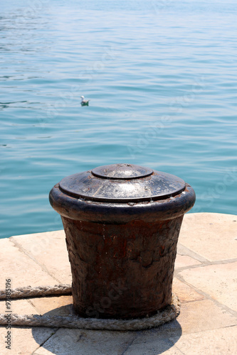 Rusty mooring bollard on a dock with nautical rope. Bright blue sea in the background. 