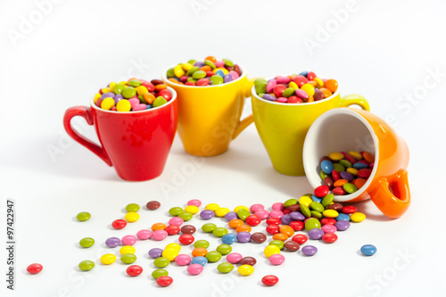 Plenty assorted color dragees in four small bright colored cups, the orange cup is felt over the sweets are spilled over the white table photo