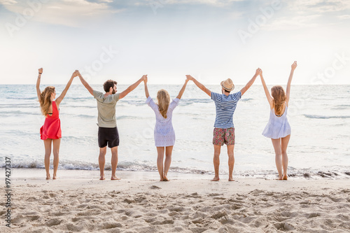 Summer beach at sunset. A group of five people holding hands and raises his arms to the sky. There are two young men and three young women during a day of rest in the sea with deserted beach