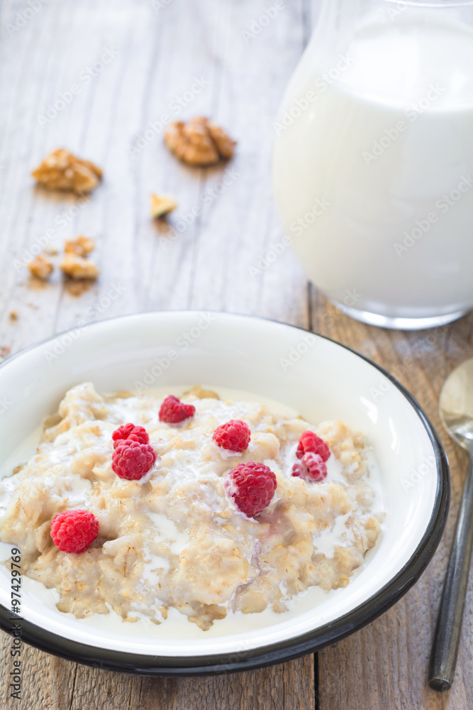 Oatmeal porridge with raspberries and milk on textured wooden table, country style healthy breakfast. Vertical composition