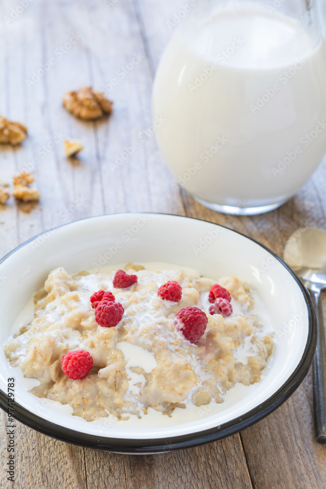 Oatmeal porridge with raspberries and milk on textured wooden table, country style healthy breakfast. Vertical composition