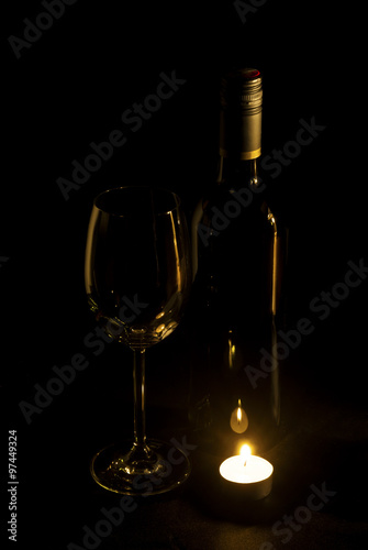 A wine glass and a bottle of wine is illuminated of candle light.