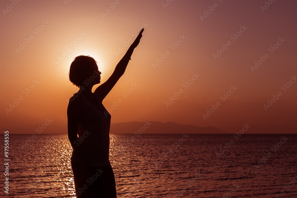 Fototapeta premium Silhouette of young girl standing on the beach with hand up, at the sunset against the sun