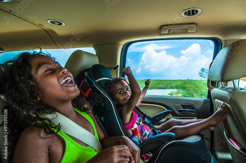 Brazilian girls singing and laughing sitting on backseats in car. Travelling female children in child safety seat with seatbelt on a summer day in tropical area