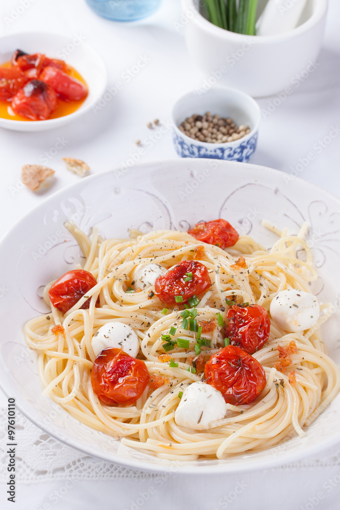 Italian spaghetti pasta with baked cherry tomatoes, mozzarella and spring onions in a white bowl on a white table.