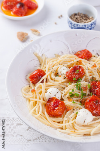 Italian spaghetti pasta with baked cherry tomatoes, mozzarella and spring onions in a white bowl on a white table.