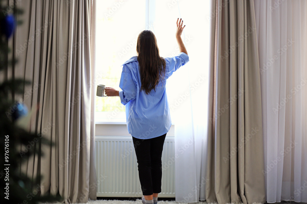 Woman with cup of coffee looking through the window in the room Woman with cup of coffee looking through the window in the room