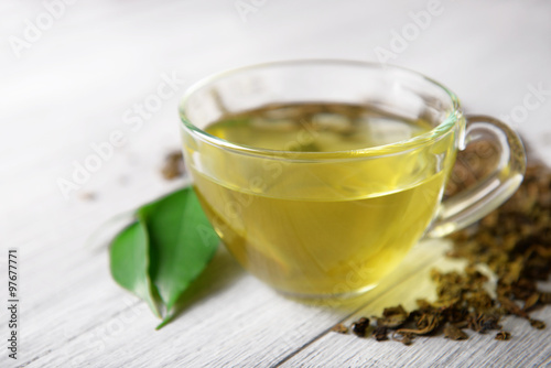 Glass cup of tea with green leaves and scattered tea around on grey wooden background, close up