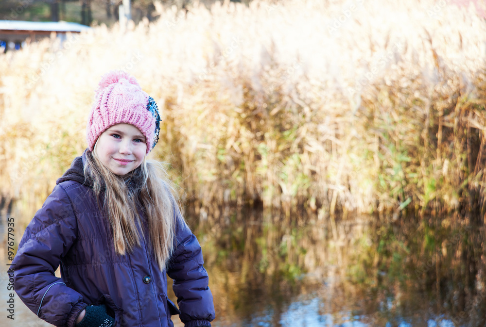 Little beautiful blonde girl in pink hat on background of autumnal lake.