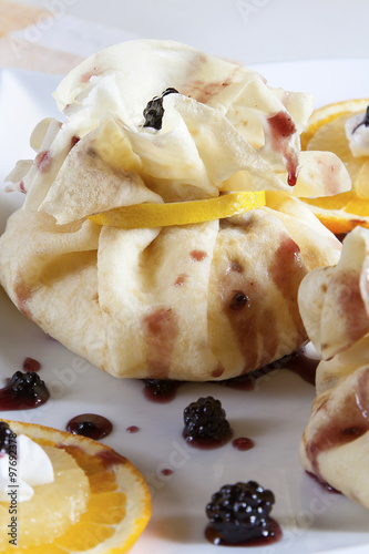 Pancake wrapped as bag, with sweet filling served on a white plate with blackberries, orange, lemon and whipped cream. 