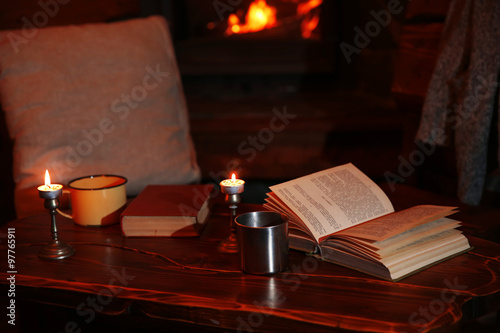 Hot tea or coffee in mug  book and candles on vintage wood table. Fireplace as background