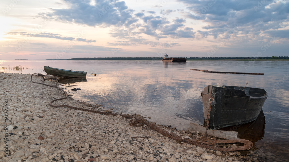 Two laid up wooden boats and cargo ship against Severnaya Dvina River panorama at sunset. Bereznik settlement, Arkangelsky region, Russia.
