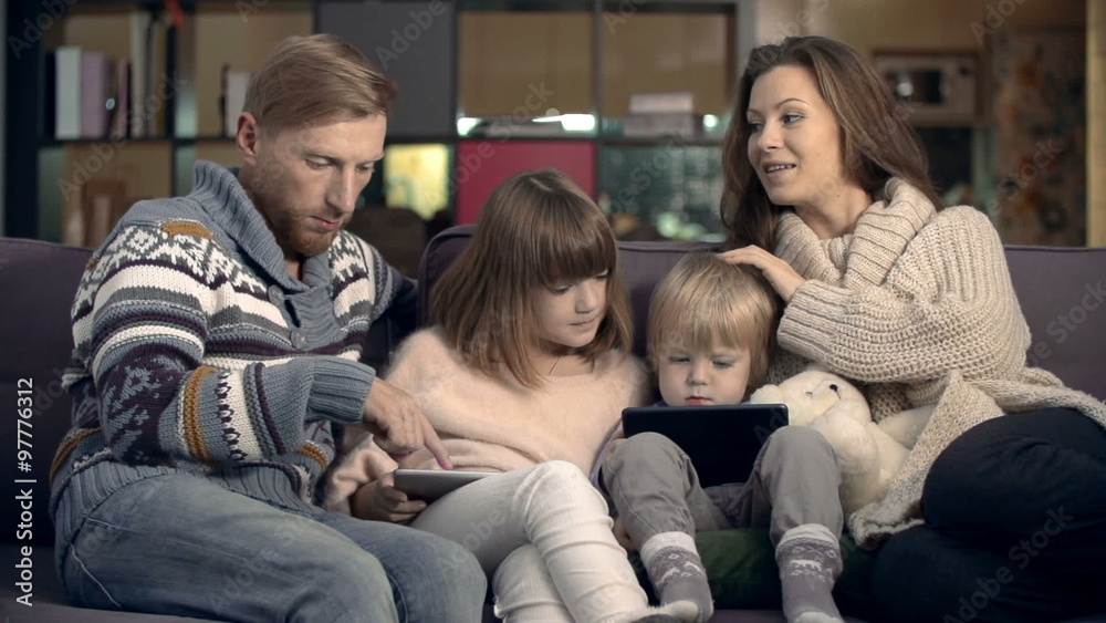 Happy family spending their evening together at home: kids playing on their tablets, mom and dad talking to each other Happy family spending their evening together at home: kids playing on their tablets, mom and dad talking to each other