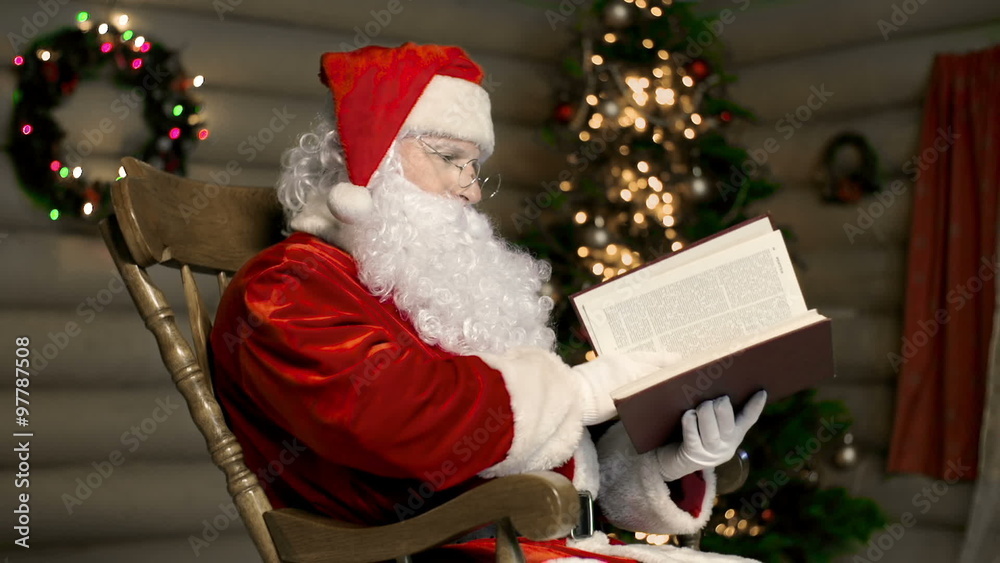 Santa sitting in wooden armchair near illuminated Christmas tree and reading a book Santa sitting in wooden armchair near illuminated Christmas tree and reading a book