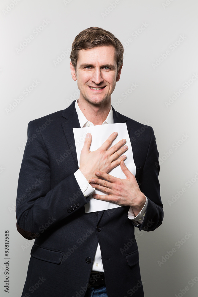 Blond-haired businessman presses a piece of paper to his chest with a cheeky grin while in front of a gradient background Blond-haired businessman presses a piece of paper to his chest with a cheeky grin while in front of a gradient background