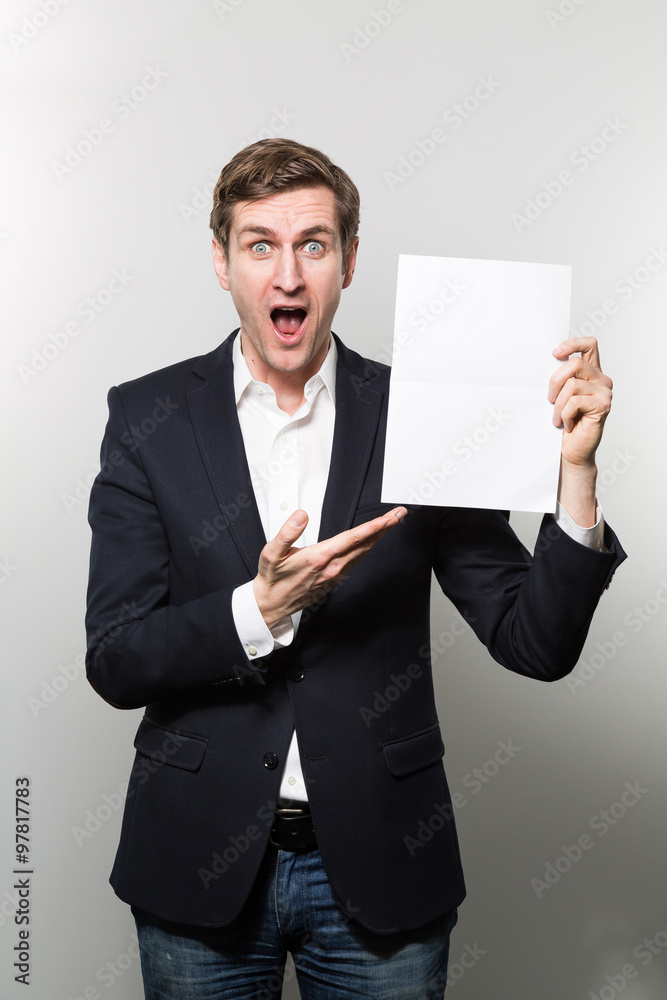 Blond-haired european businessman shows something on a piece of paper with a incredible face while in front of a gradient background Blond-haired european businessman shows something on a piece of paper with a incredible face while in front of a gradient background