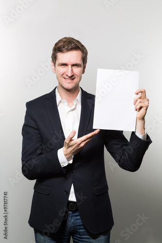 Blond-haired european businessman shows something on a piece of paper with a cheeky grin while in front of a gradient background © Nektarstock
