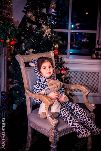Pretty little girl smiling with teddy bear near the Christmas tree sitting in vintage chair. Happy New Year.
