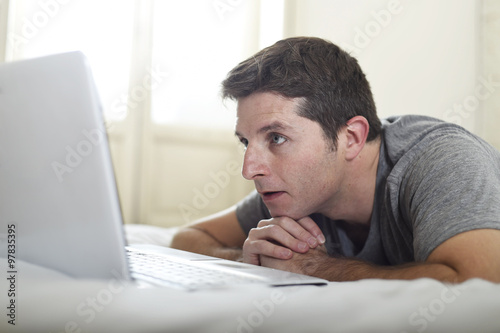portrait of young attractive man lying on bed watching on line movie in computer laptop at home in internet concept