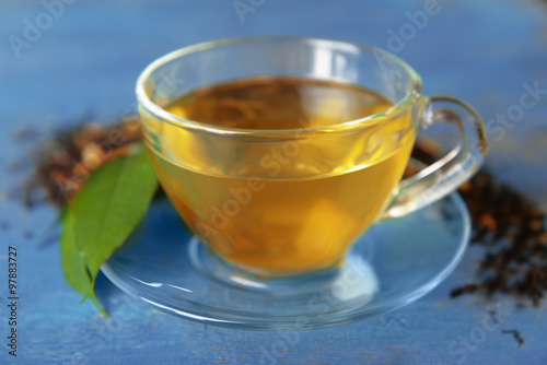 Glass cup of tea with green leaves and scattered tea around on blue wooden background